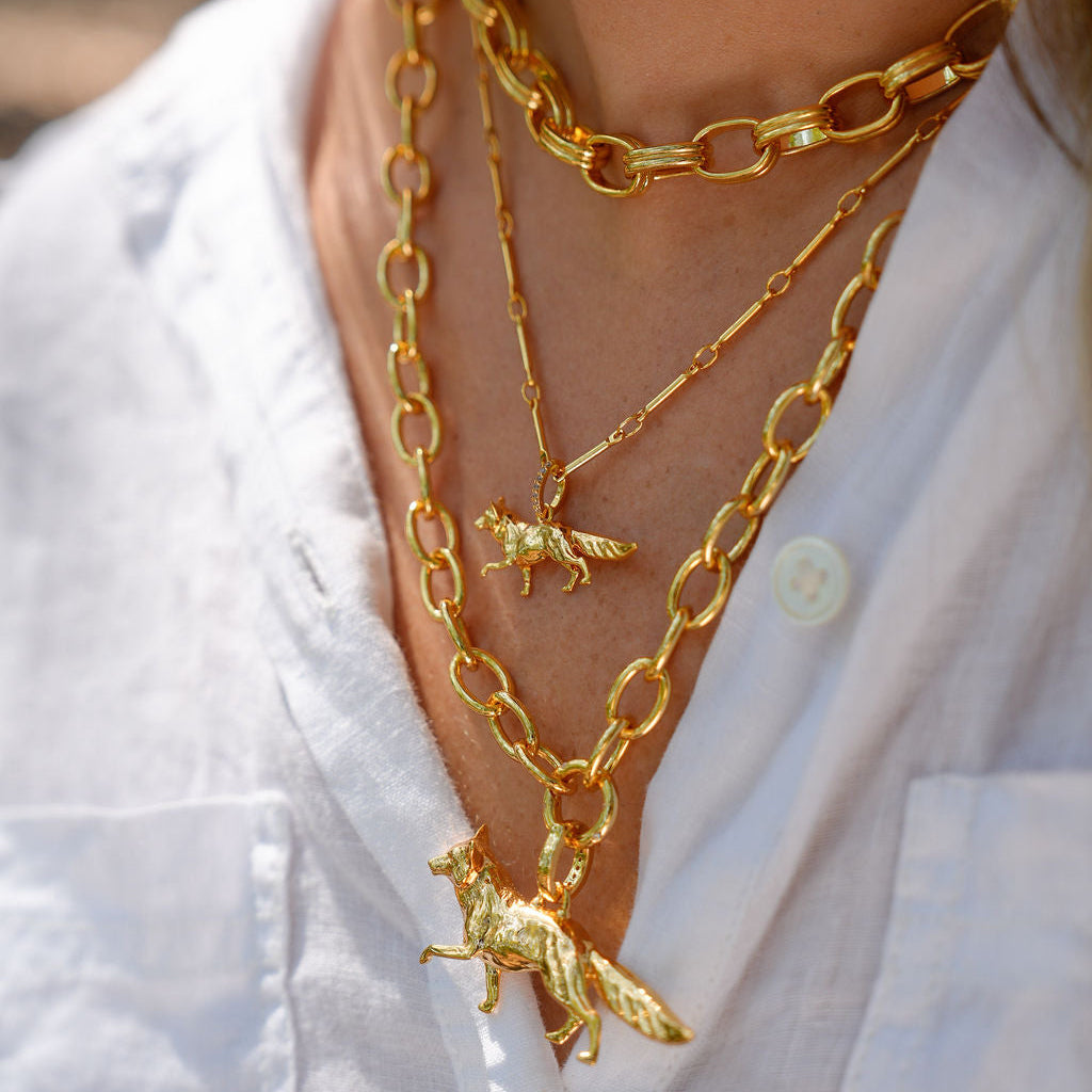 A close-up of a person wearing layered gold necklaces, including a chunky chain, a delicate chain with a small fox pendant, and a longer chain with a larger fox pendant, styled with a white linen shirt.