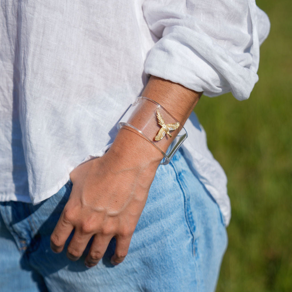 A person outdoors wearing a clear rectangular cuff bracelet with a small gold butterfly or wing-like charm on the wrist, paired with a white shirt and blue jeans.