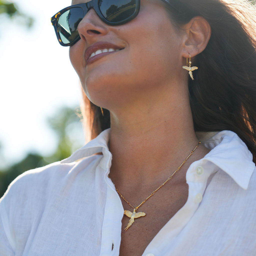 A woman wearing a white linen button-up shirt, gold shark tooth necklace, matching gold shark tooth earrings, and dark sunglasses, captured in bright outdoor sunlight.