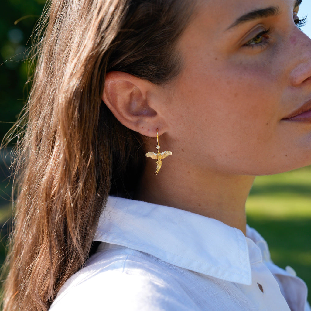 A close-up side profile of a woman wearing a delicate gold dangle earring shaped like a small bird or flower, paired with a white collared shirt against a blurred green outdoor background.