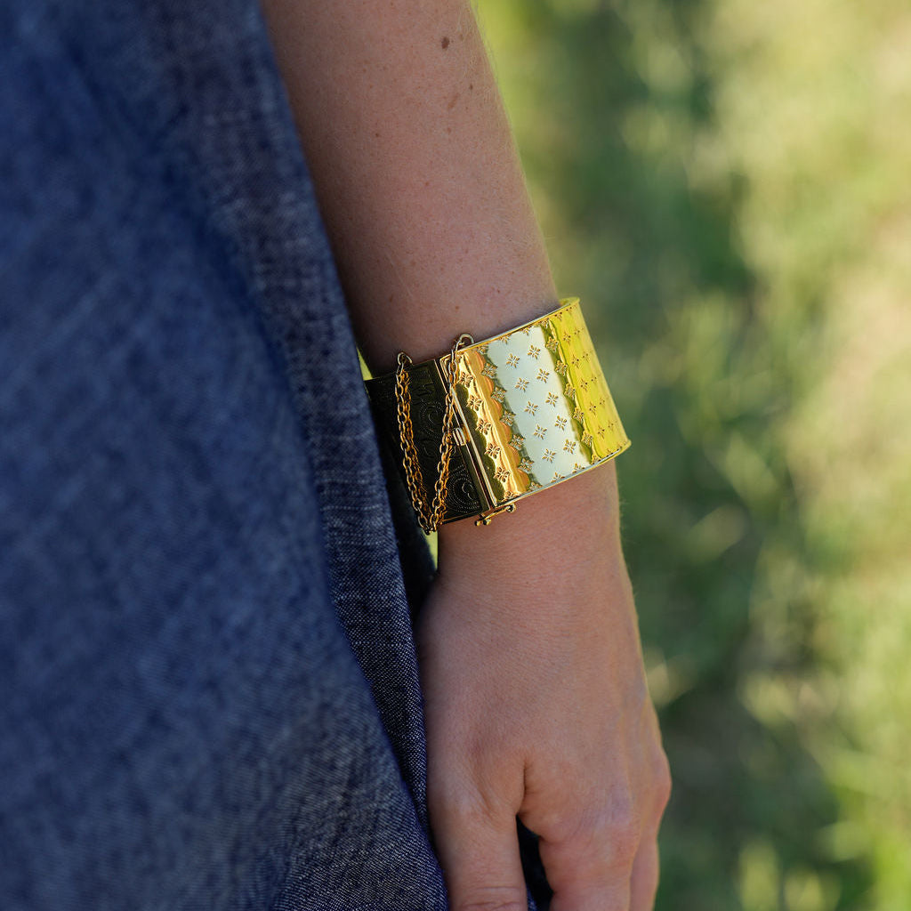 A close-up of a person's hand and forearm wearing a wide, chunky gold cuff bracelet with textured panels, set against a blurred outdoor background.