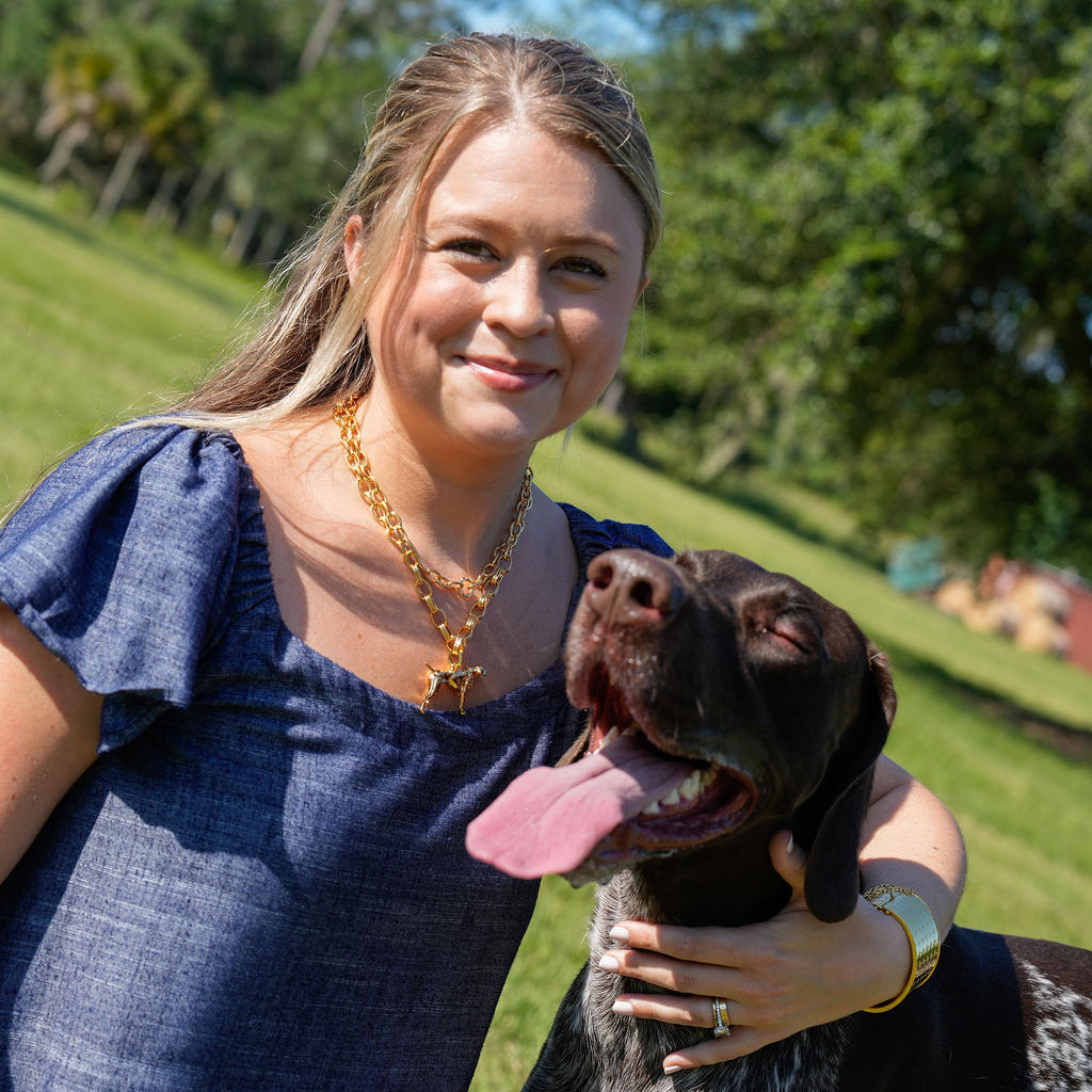 A woman in a blue dress smiling outdoors next to a black and white spotted dog, with green grass and trees in the background.