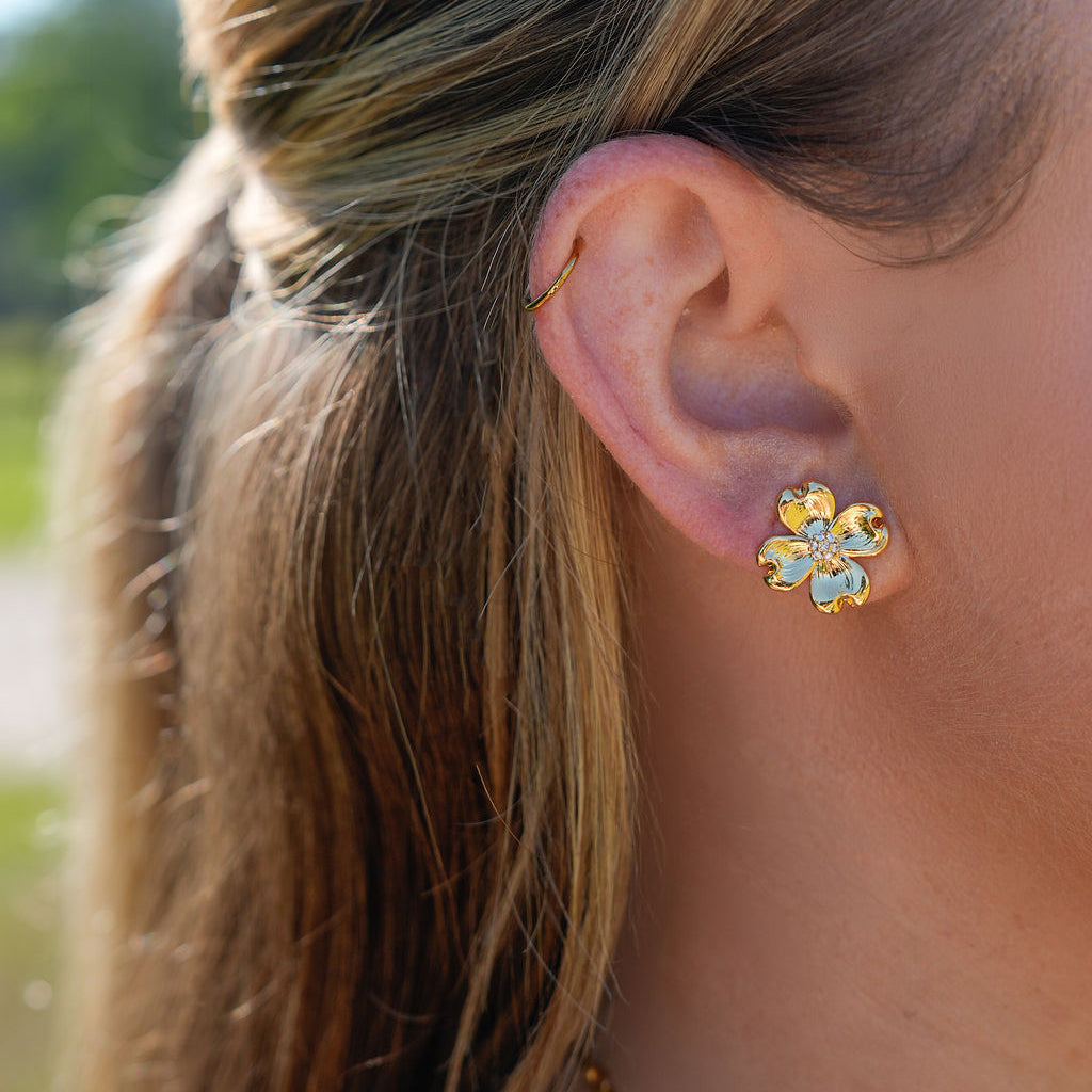 A close-up side profile of a woman's ear wearing a small gold flower-shaped stud earring with clear rhinestones, against a softly blurred outdoor background.