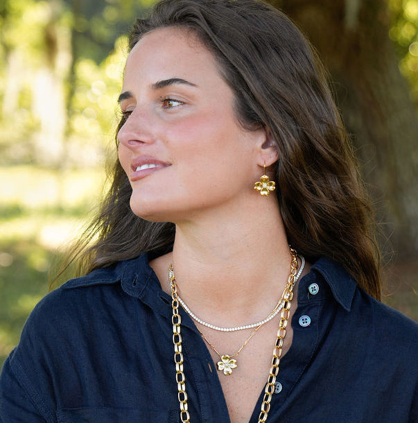 A woman sitting on a park bench wearing a navy blue button-up shirt, layered gold necklaces, gold bracelets, and a gold earring, with a blurred outdoor background of trees and grass.