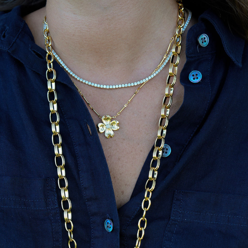 A close-up of a person wearing layered necklaces over a dark navy button-up shirt. The necklaces include a thick gold chain, a delicate gold flower pendant necklace, a thin gold chain, and a short pearl-like beaded necklace.