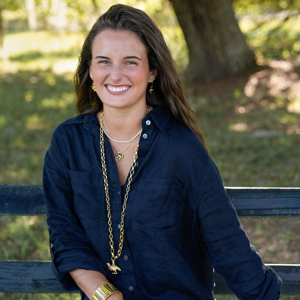 A smiling woman with long brown hair sits on a dark wooden park bench outdoors. She is wearing a navy blue button-up shirt, light khaki shorts, layered gold necklaces, and gold bracelets, with a blurred green and brown wooded background.