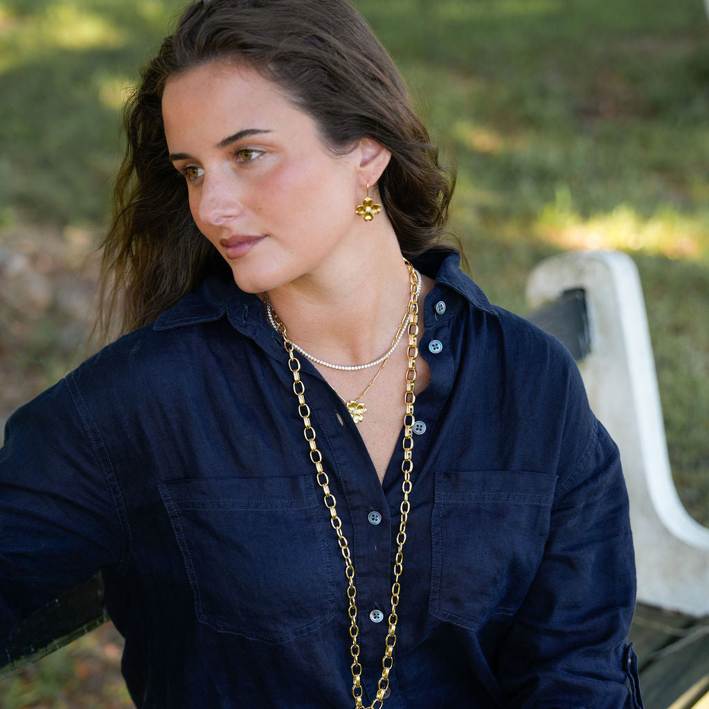 A woman wearing a navy blue button-up shirt and layered gold necklaces sits on a bench in an outdoor park setting.
