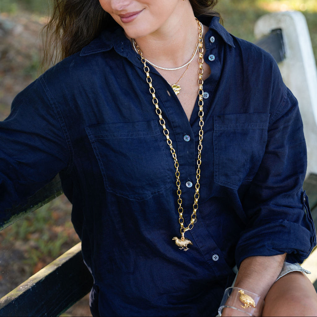 A woman wearing a navy blue button-up shirt and layered gold necklaces, sitting outdoors on a bench with a blurred green background.