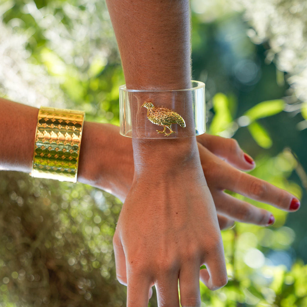 A clear cylindrical cuff bracelet with a gold-toned bird charm, shown worn on a wrist outdoors with another gold bracelet visible on another wrist, against a blurred green foliage background.