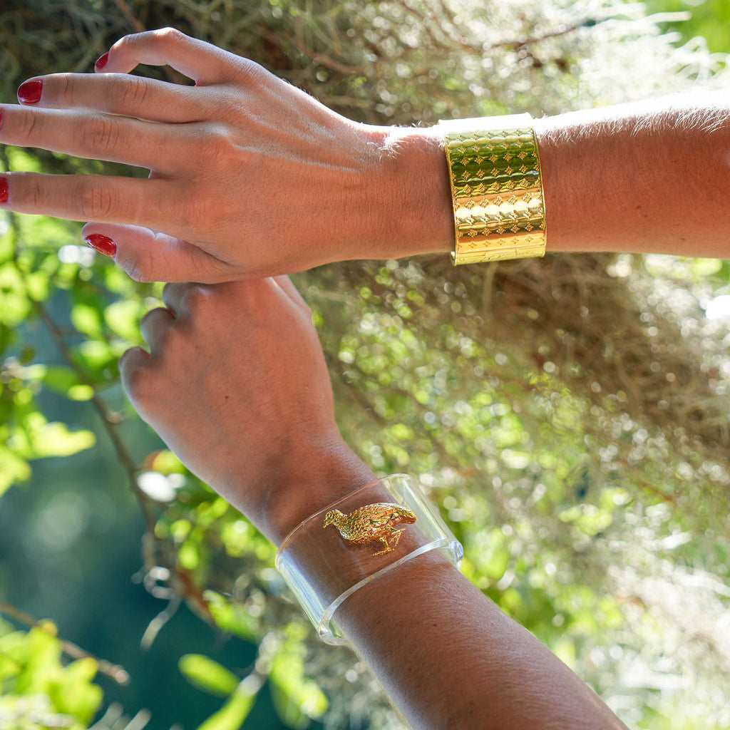 A close-up lifestyle shot of a person's hands and forearms wearing two gold bracelets against a natural background of green foliage and hanging moss. One bracelet is a wide, textured gold cuff, and the other is a clear cuff featuring a gold leaf charm.
