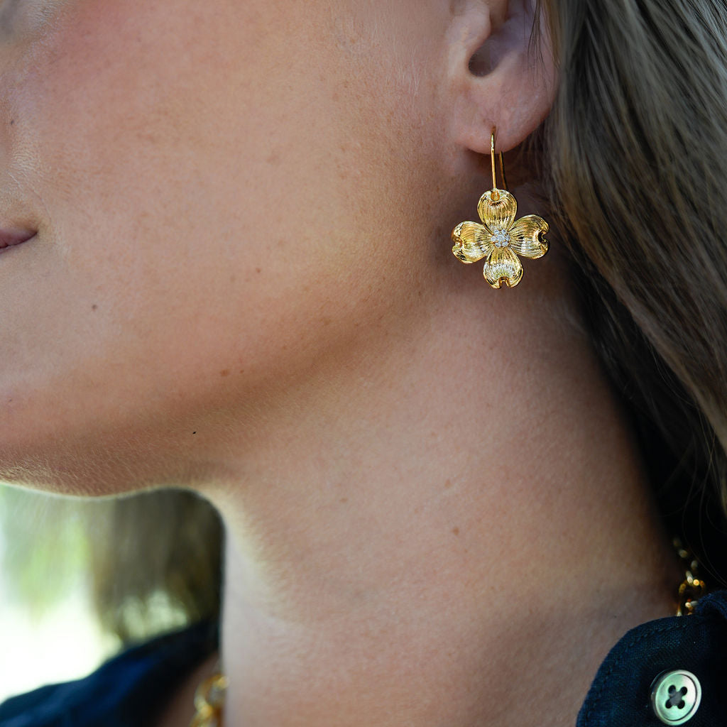 A close-up lifestyle shot of a model wearing a gold four-leaf clover dangle earring, layered with gold and silver necklaces, paired with a dark navy button-up shirt.