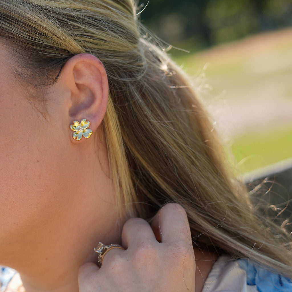A close-up lifestyle shot of a woman wearing a gold flower stud earring with clear rhinestones, along with a gold cross pendant necklace and a matching gold flower bracelet.
