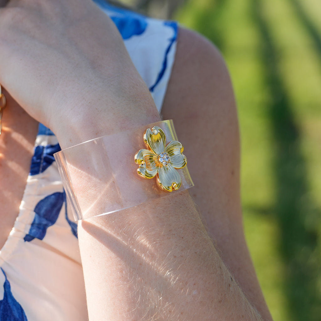A close-up of a woman's arm wearing a wide, clear plastic cuff bracelet with a gold and white flower charm, set against a blurred green outdoor background.