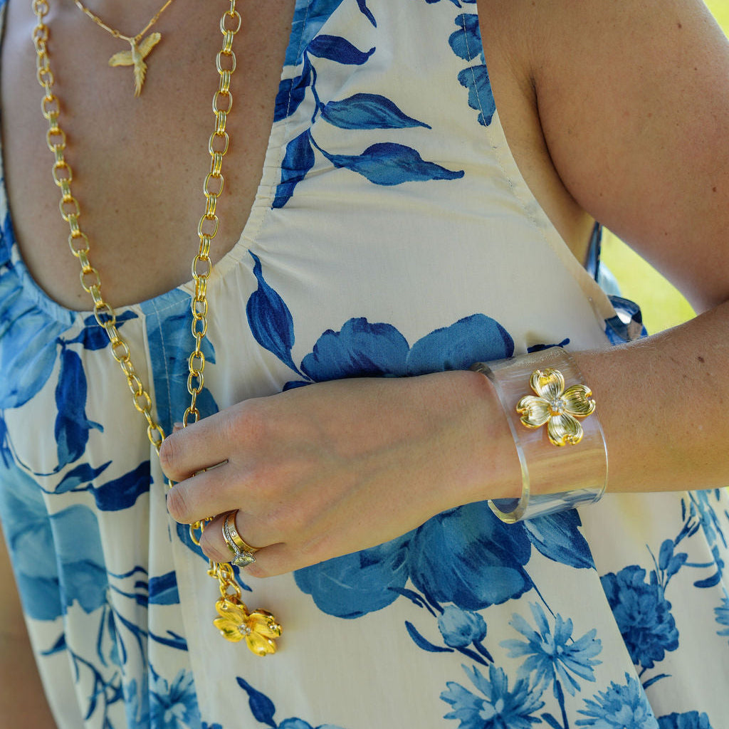 A close-up lifestyle shot of a person wearing a white sleeveless dress with a blue floral print, accessorized with gold necklaces, a flower-shaped bracelet, and a clear cuff bracelet.