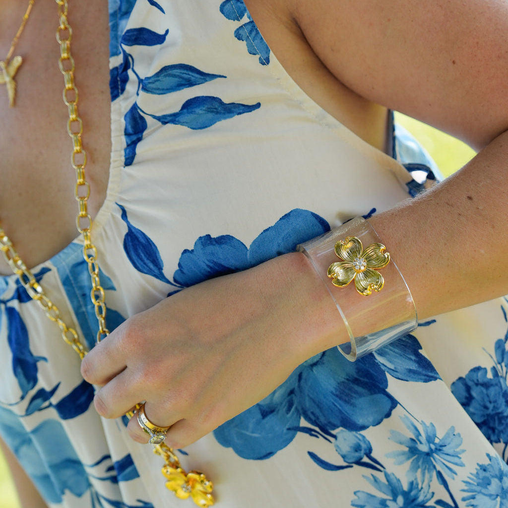 A close-up lifestyle shot of a person wearing a white sleeveless dress with a blue floral print, accessorized with gold jewelry including a necklace, ring, and clear bracelet.