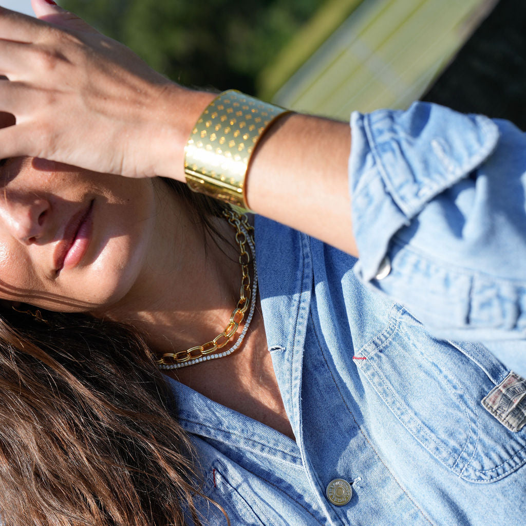 A close-up lifestyle shot of a woman wearing a wide, textured gold cuff bracelet on her wrist, a gold chain necklace, and a light blue denim shirt.