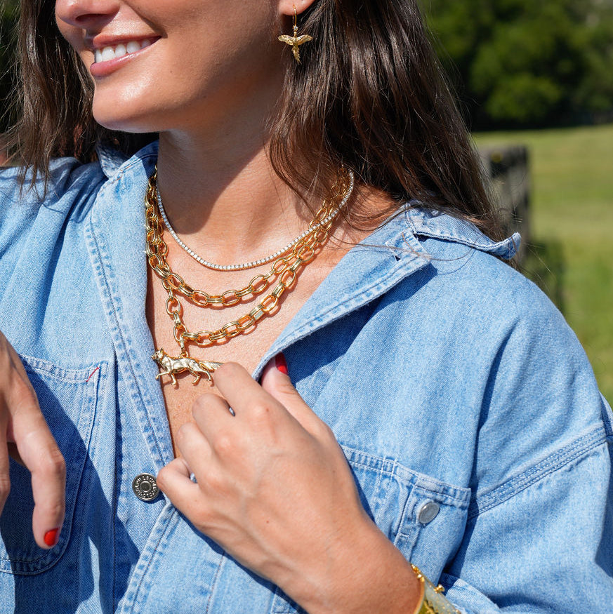 A woman outdoors wearing layered gold necklaces, a gold cuff bracelet, and a gold cross earring, paired with a light blue denim shirt against a green grassy background.