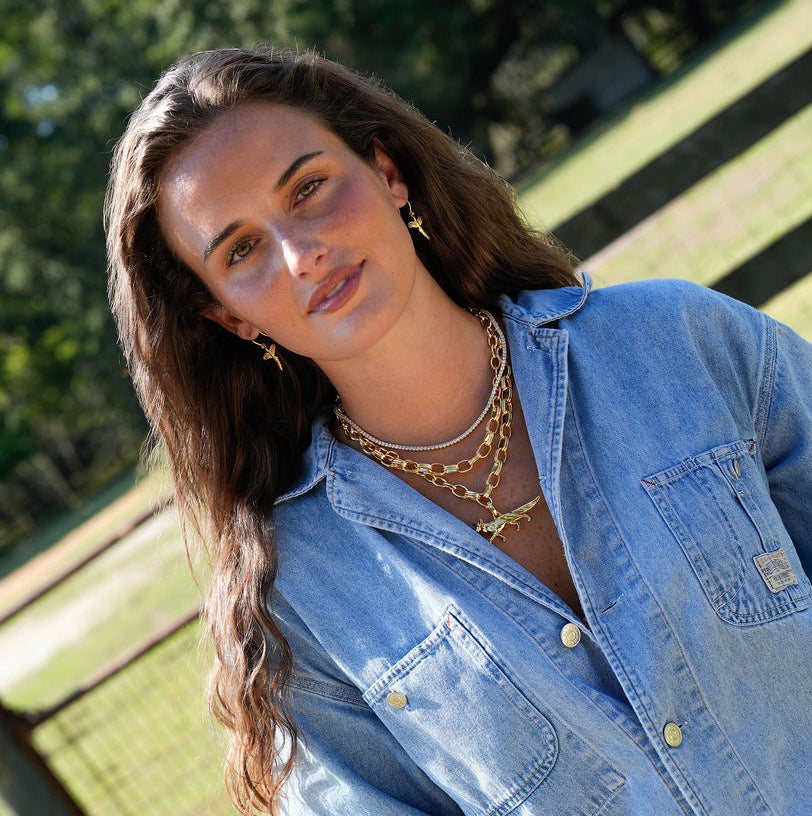A woman wearing a light blue denim button-up shirt and white pants, standing outdoors in a grassy field with a wooden fence in the background.