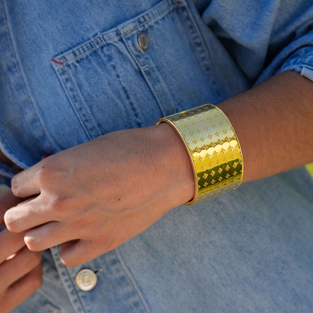 A close-up of a person wearing a wide, chunky gold cuff bracelet with a textured chevron pattern, styled with a blue denim shirt.