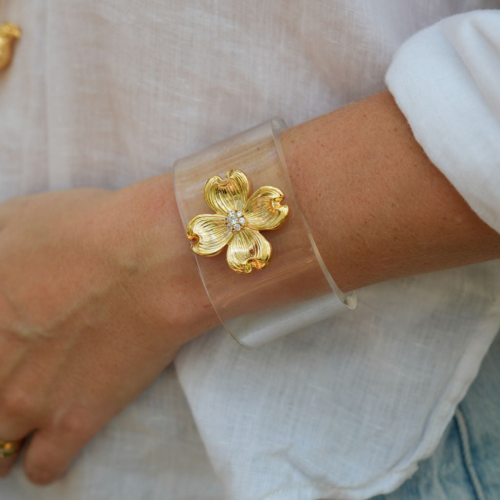 A close-up of a person's wrist wearing a clear cuff bracelet with a gold flower-shaped charm in the center, paired with a white linen top.