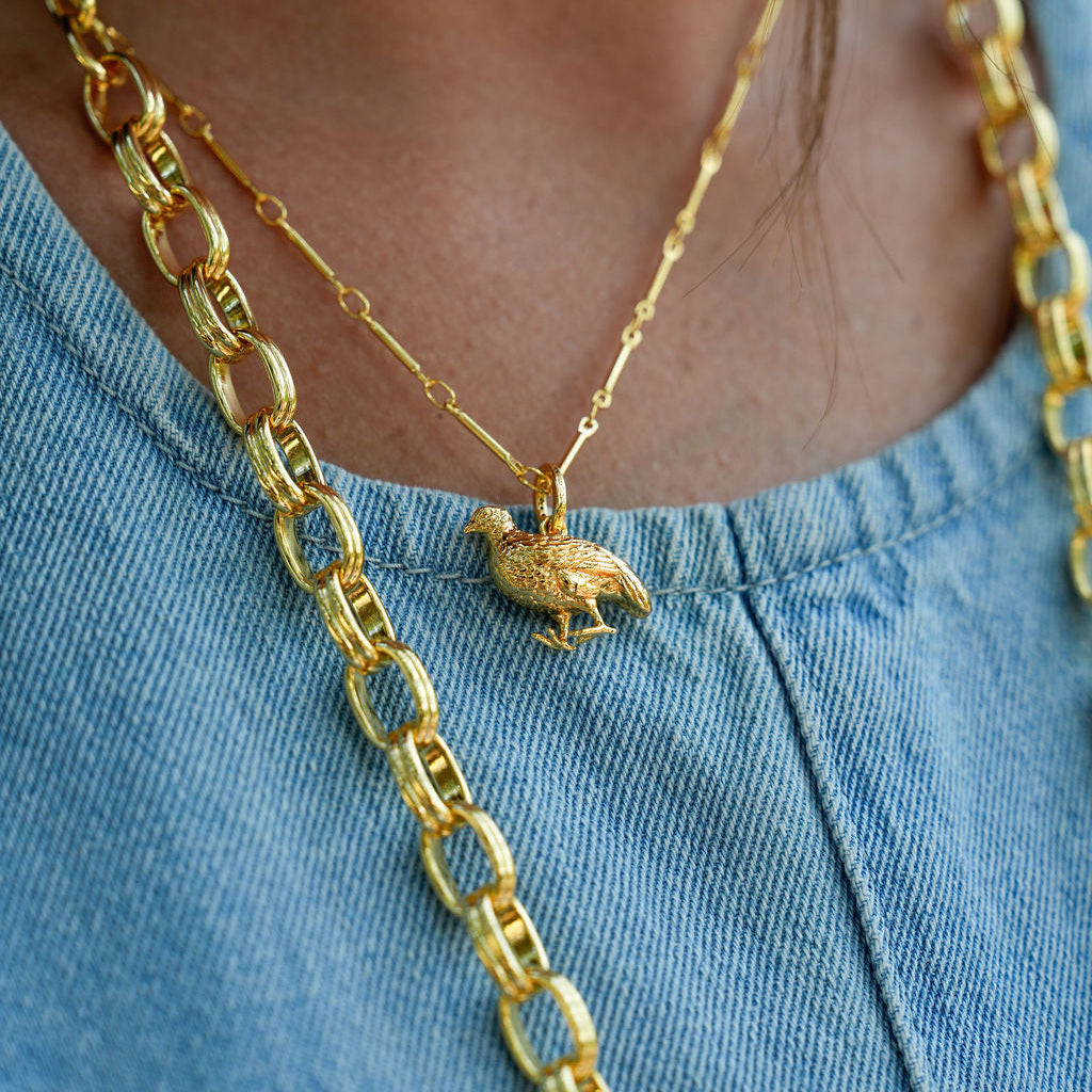 A close-up of a person wearing layered gold necklaces over a light blue denim top. One necklace has a small bird pendant, and the other is a chunky link chain.