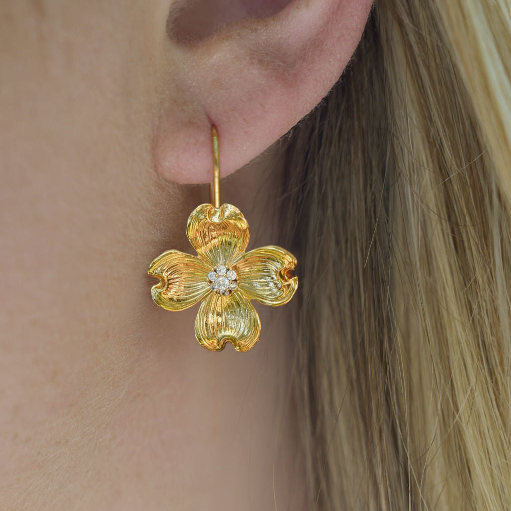 A close-up of a person's ear wearing a textured gold flower-shaped dangle earring with a small clear stone in the center.