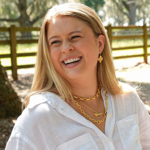 A woman smiling outdoors wearing a loose white button-up shirt and light denim shorts, standing in a sunlit wooded area with trees and a fence in the background.
