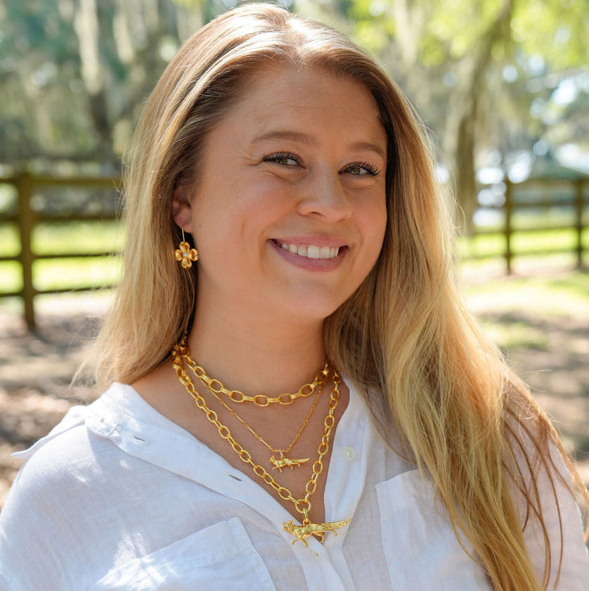 A smiling woman with long blonde hair wearing layered gold necklaces and earrings, posing outdoors in a white button-up shirt against a blurred background of trees and a fence.