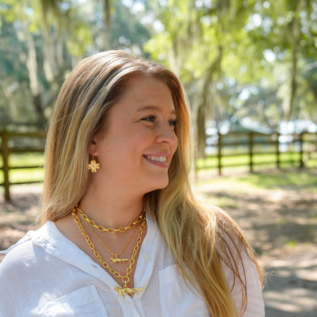 A smiling blonde woman wearing layered gold necklaces and a gold cross pendant, styled with a white button-up shirt, standing outdoors in a sunny park with trees and a fence in the background.