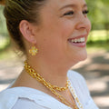 A woman in a white blouse wearing gold jewelry, including a gold flower-shaped earring and layered gold chain necklaces, photographed outdoors with a blurred green background.
