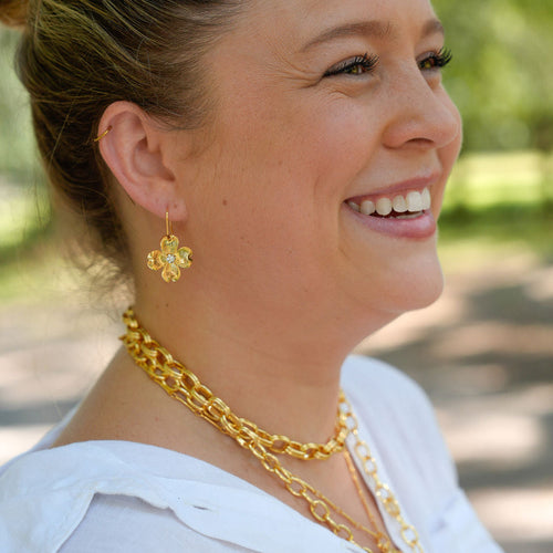 A woman in a white blouse wearing gold jewelry, including a gold flower-shaped earring and layered gold chain necklaces, photographed outdoors with a blurred green background.