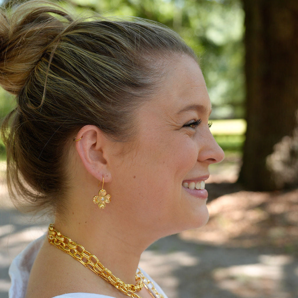 A woman in profile wearing a chunky gold chain necklace and matching gold flower-shaped earrings, dressed in a white top against a blurred outdoor background.