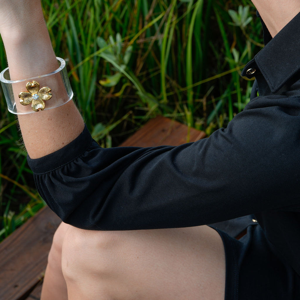 A woman wearing a wide clear cuff bracelet with a gold floral design on her wrist, photographed outdoors in front of green grass.