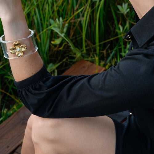 A woman wearing a wide clear cuff bracelet with a gold floral design on her wrist, photographed outdoors in front of green grass.