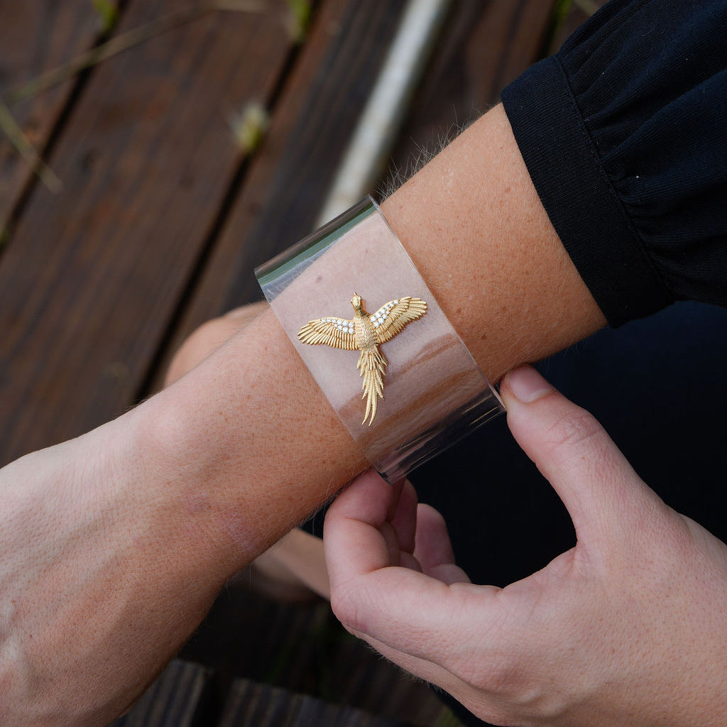 A close-up of a person's forearm wearing a clear cuff bracelet with a gold bird pendant, set against a dark wooden deck background.