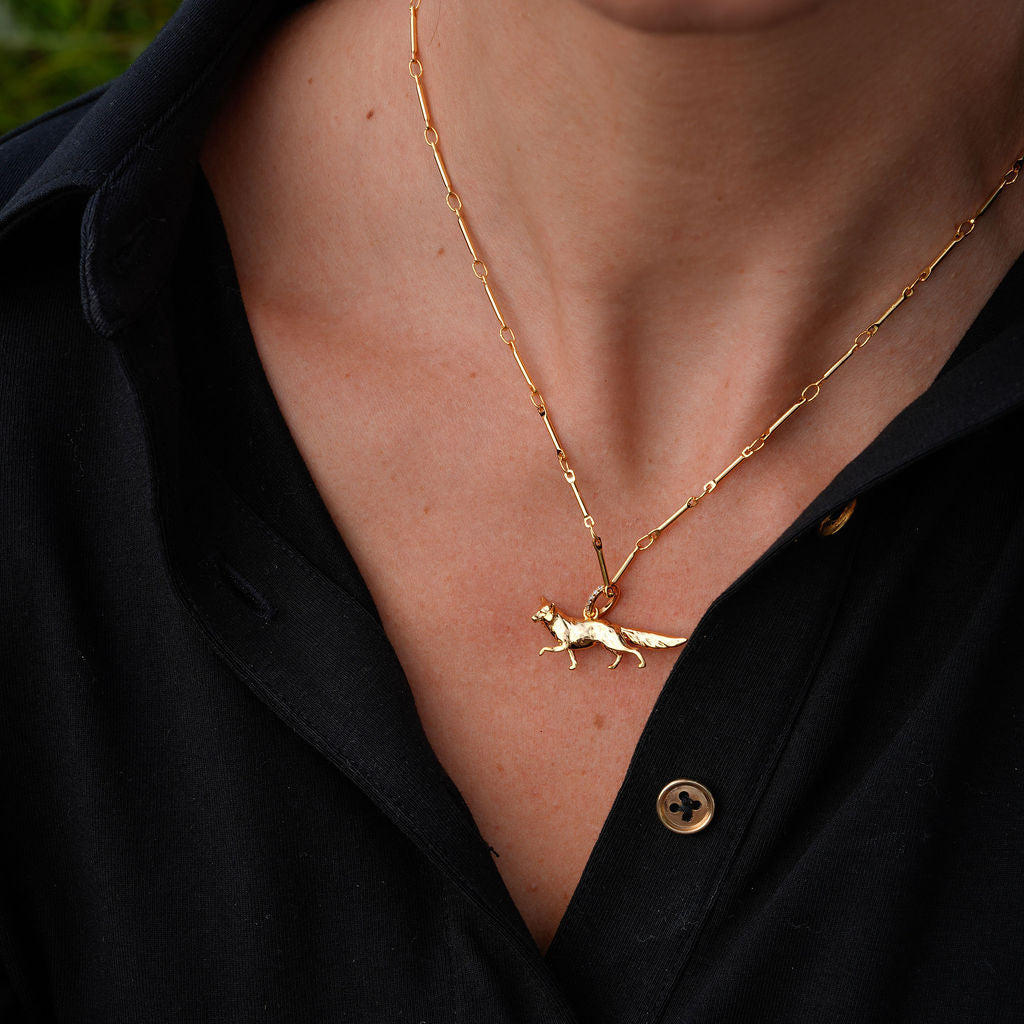 A close-up of a person wearing a delicate gold chain necklace with a small running fox pendant, styled with a black button-down shirt.