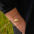 A close-up of a person's forearm wearing a clear adhesive bandage with a small gold bird charm attached, outdoors with grass in the background.