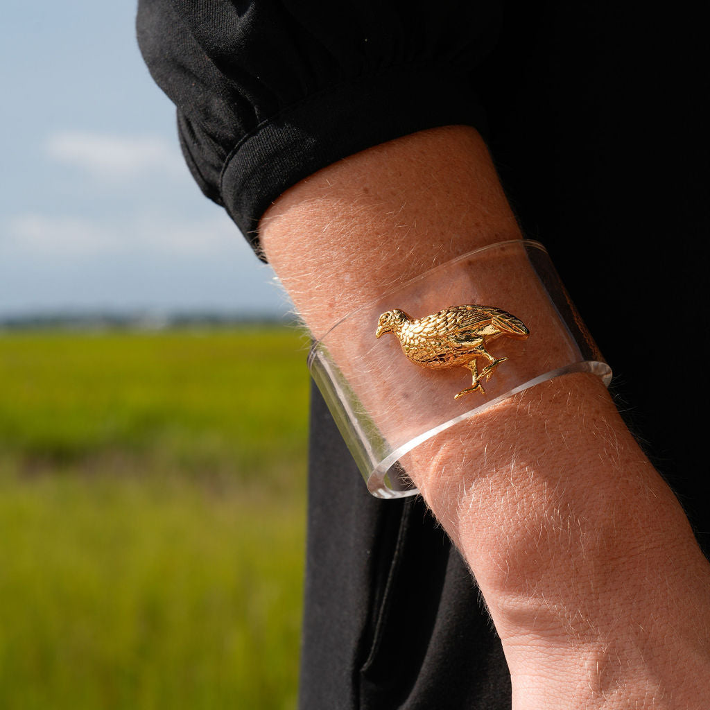 A person wearing a clear cuff bracelet with a detailed gold bird charm, photographed outdoors against a blurred green field background.
