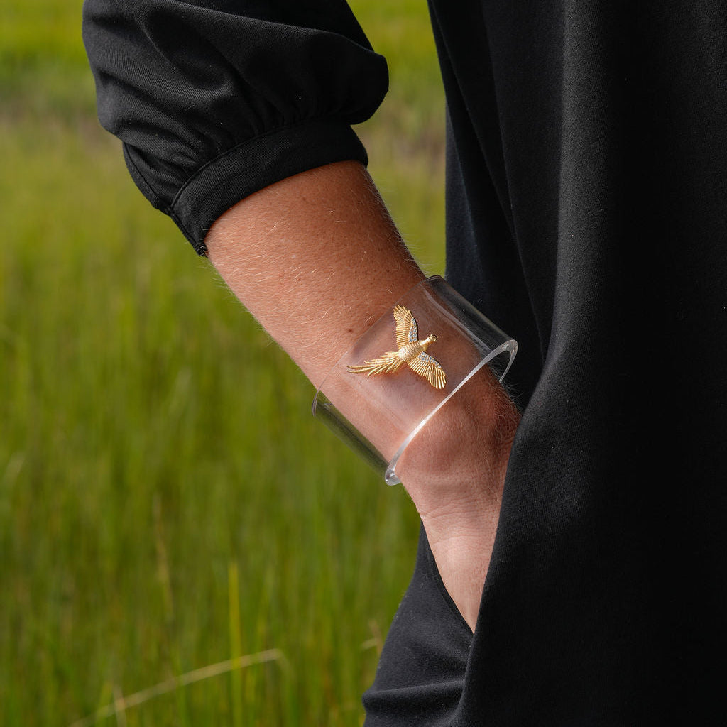 A person wearing a clear cuff bracelet with a small gold butterfly charm, photographed outdoors in a grassy field.