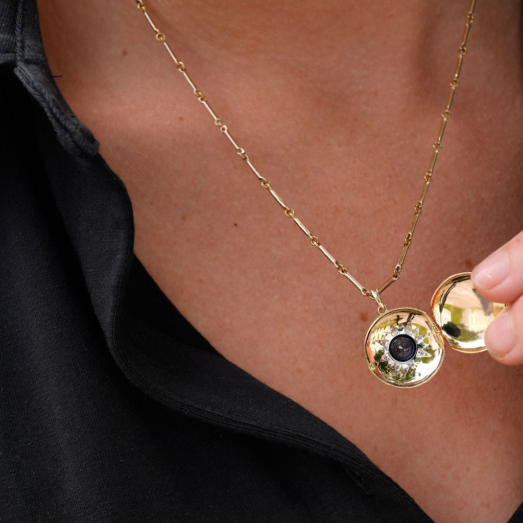 A close-up of a person wearing a delicate gold chain necklace with a round locket pendant. The locket is open, revealing a small black evil eye motif in the center, and a hand is holding the other side of the locket.
