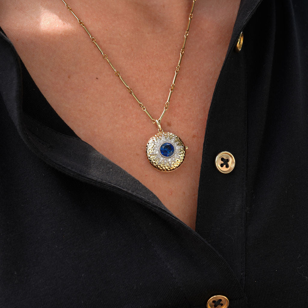 A close-up of a person wearing a gold chain necklace with a circular pendant featuring a blue evil eye design surrounded by small clear stones, paired with a black button-up shirt.