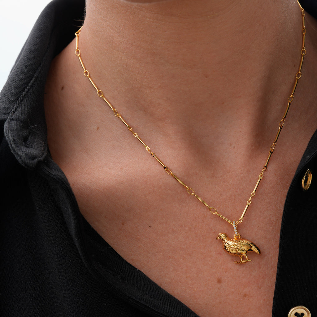 A close-up of a person wearing a delicate gold chain necklace with a small gold bird pendant, styled with a black button-down shirt.