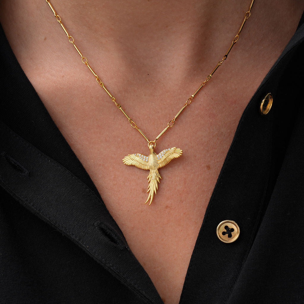 A close-up of a person wearing a gold chain necklace with a bird pendant, styled with a black button-down shirt.