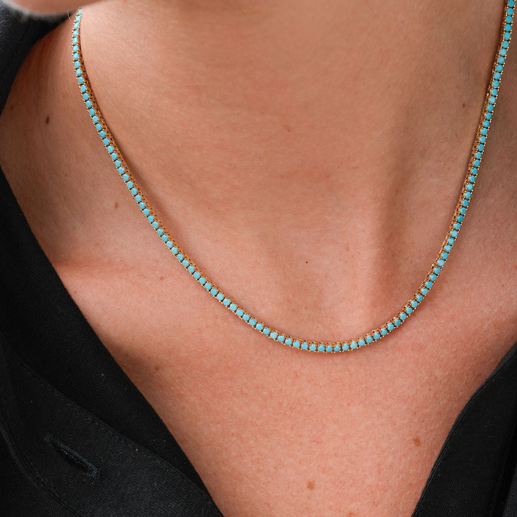 A close-up of a person wearing a delicate gold chain necklace with small turquoise-blue stones, paired with a black collared shirt.