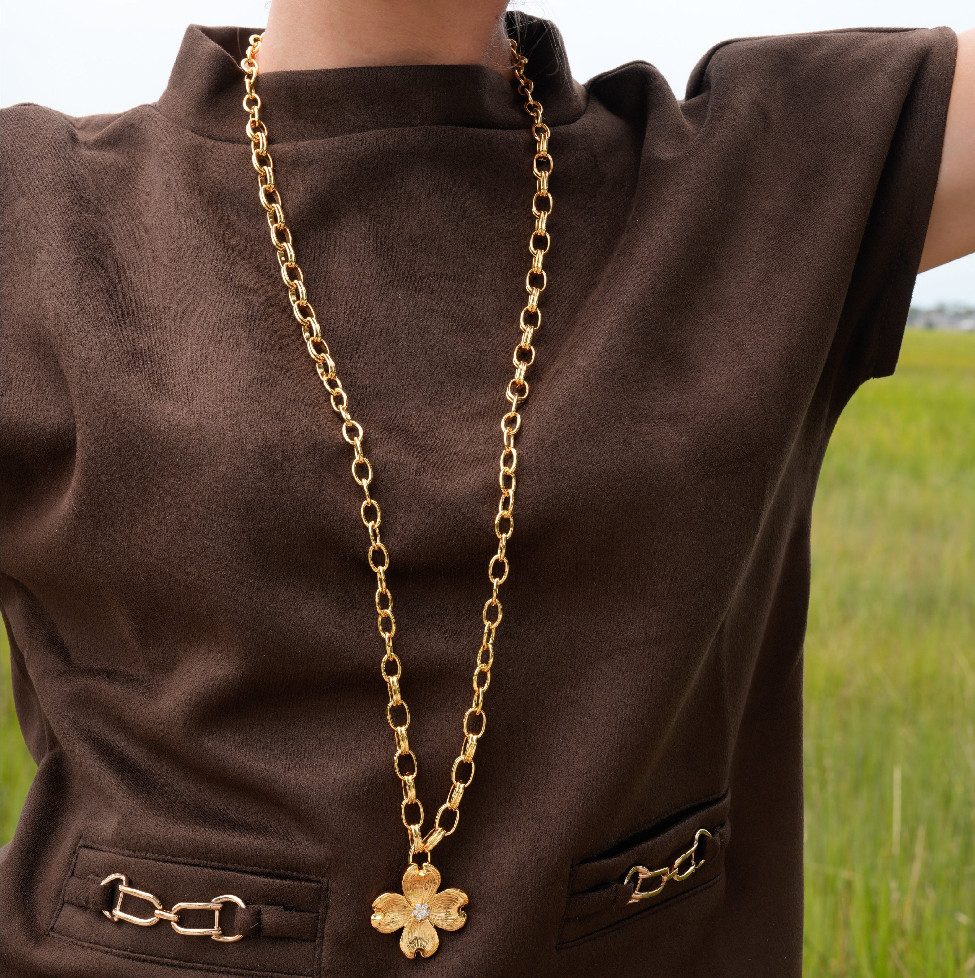 A gold-tone floral pendant necklace with a small clear stone in the center, worn over a brown sleeveless top. The background shows a blurred outdoor grassy field.