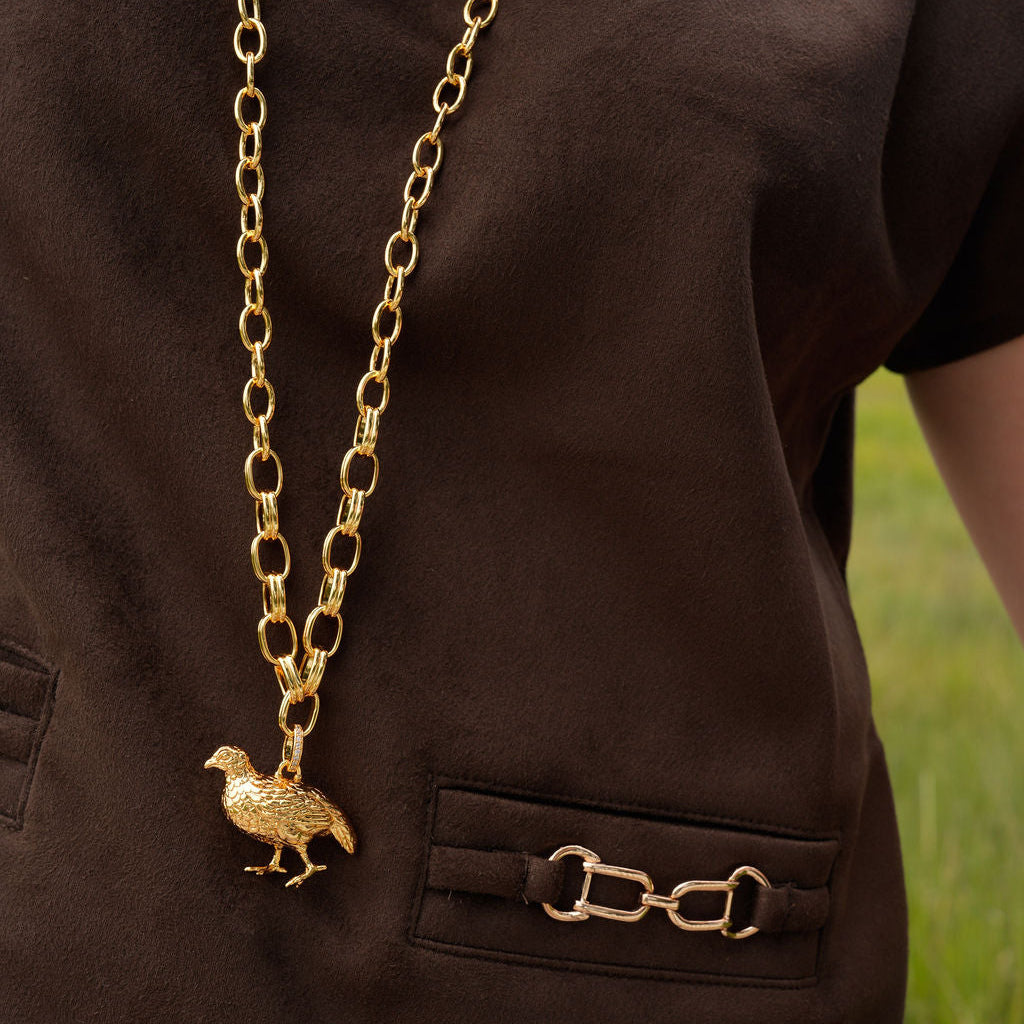 A gold chain necklace with a small bird pendant, worn over a dark brown short-sleeve shirt with a pocket and a horsebit detail on the chest.