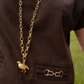 A gold chain necklace with a small bird pendant, worn over a dark brown short-sleeve shirt with a pocket and a horsebit detail on the chest.