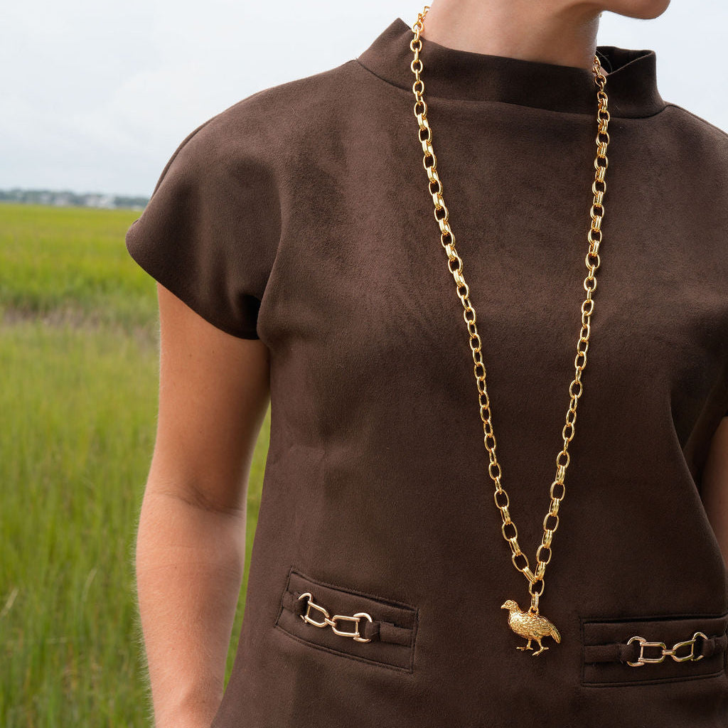 A woman wearing a brown short-sleeve top with decorative gold-tone hardware and a matching gold chain necklace with a horse pendant, photographed outdoors in a grassy field.