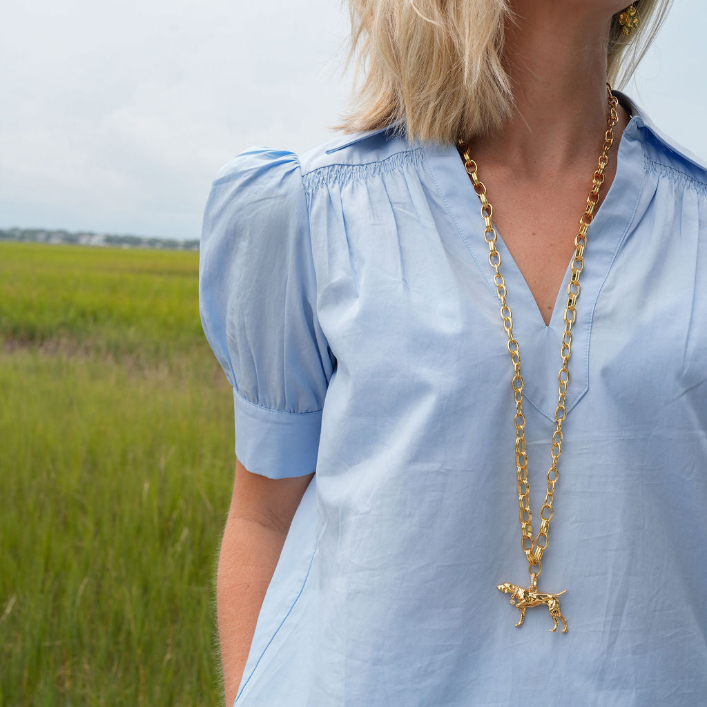 A woman wearing a light blue short-sleeve blouse with puff sleeves and a V-neck, paired with cream pants and a gold necklace, standing in a grassy field.