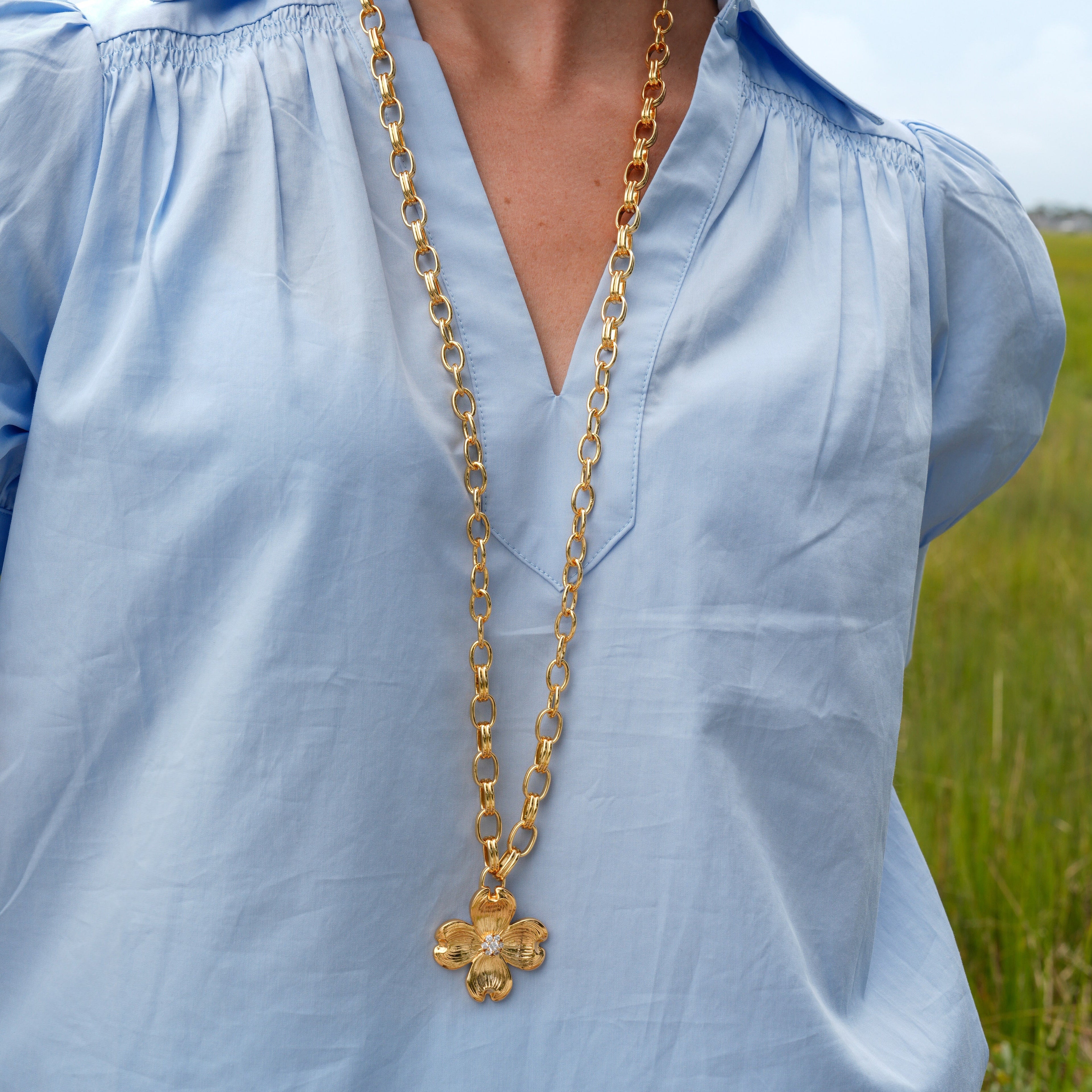 A woman wearing a light blue blouse and a long gold chain necklace with a clover-shaped pendant, photographed outdoors with a blurred green field and sky in the background.